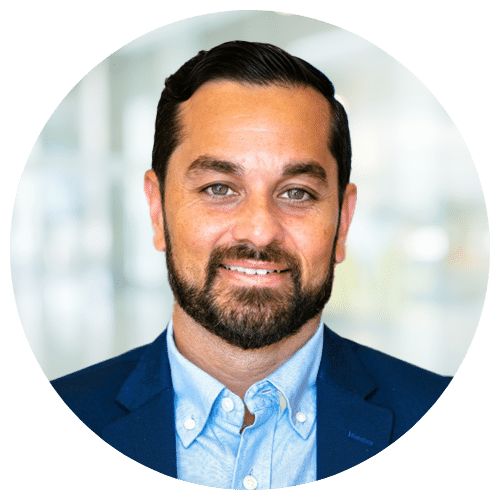Man with dark hair and beard wearing a blue suit jacket and light blue shirt, smiling at the camera with a blurred office background.
