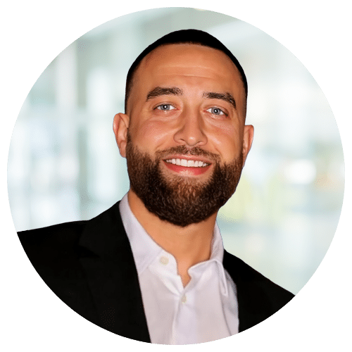 Adam Beers, National Recruiter at Jackson Nurse Professionals, with short dark hair and a trimmed beard, smiles in a black suit jacket and white shirt before a blurred indoor background.