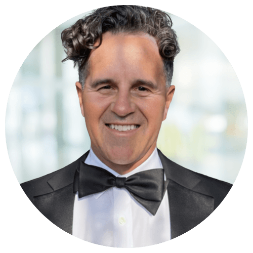 Shenan Moore, National Recruiter at Jackson Nurse Professionals, smiles in a black tuxedo and bow tie, his short curly dark hair set against a blurred light background.
