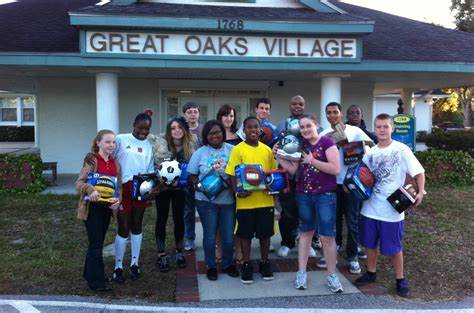 A group of people stand in front of a building labeled Great Oaks Village, giving back and creating community impact with various sports balls and equipment.
