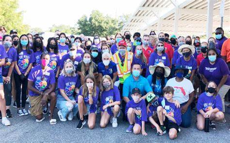 A large group of people, many wearing purple shirts and face masks, pose together outdoors under a partially covered area, showcasing their commitment to giving back and community impact.