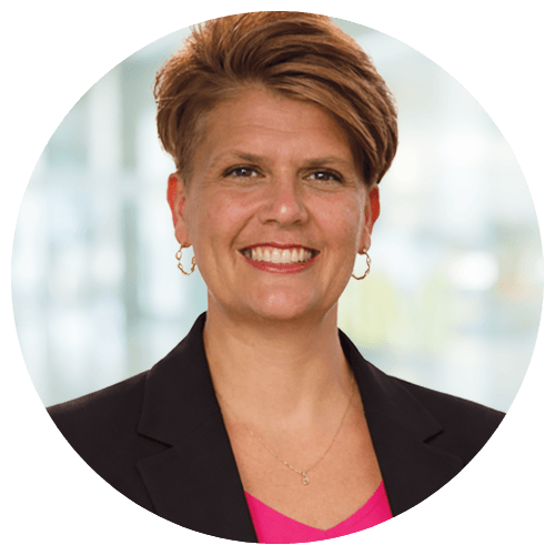 Smiling person with short light brown hair wearing a black blazer and pink top, standing in front of a blurred indoor background.