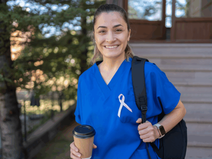 Smiling person in blue scrubs, wearing a pink ribbon, holds a coffee cup and backpack, standing outdoors—radiating potential in front of building steps.