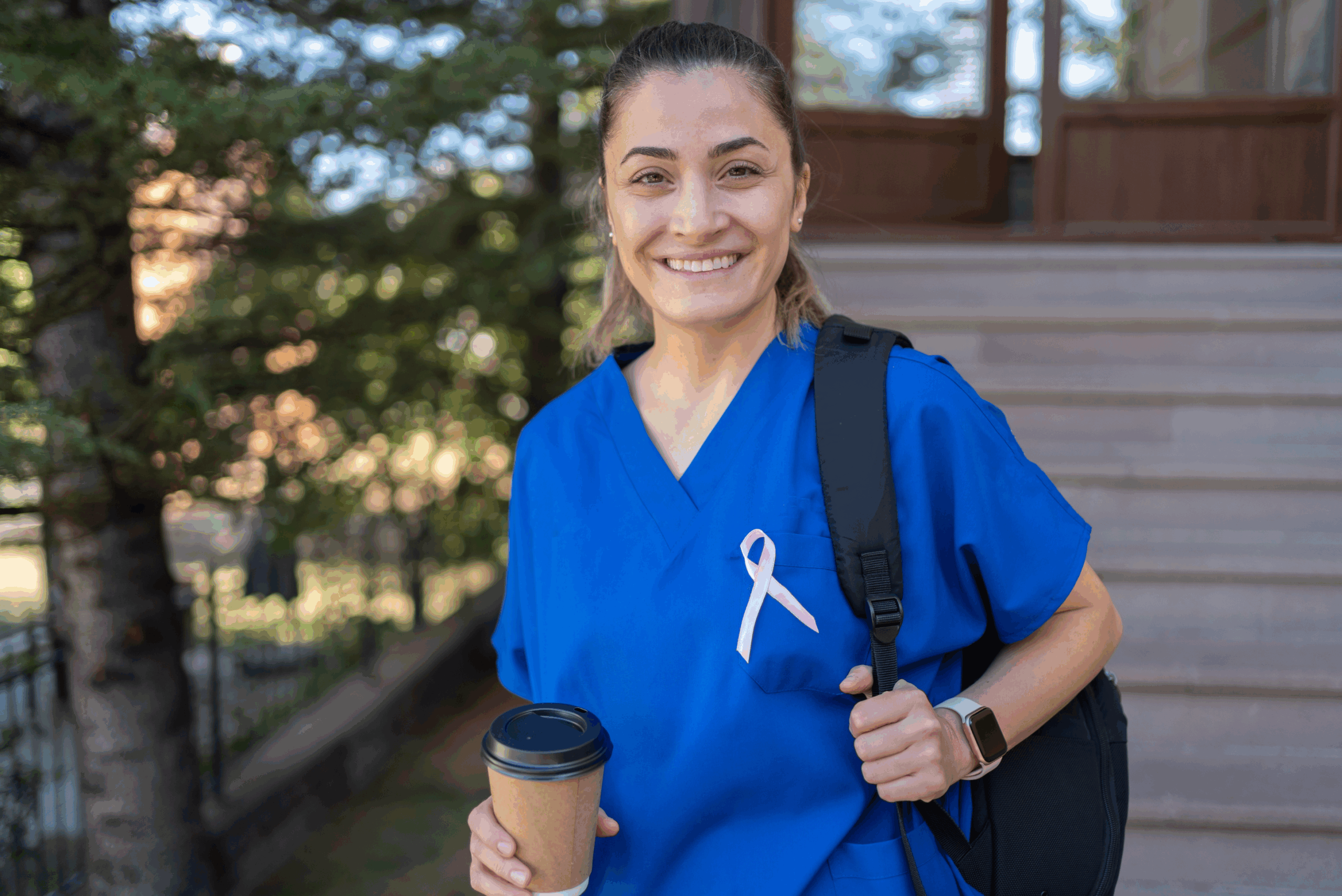 Smiling person in blue scrubs, wearing a pink ribbon, holds a coffee cup and backpack, standing outdoors—radiating potential in front of building steps.
