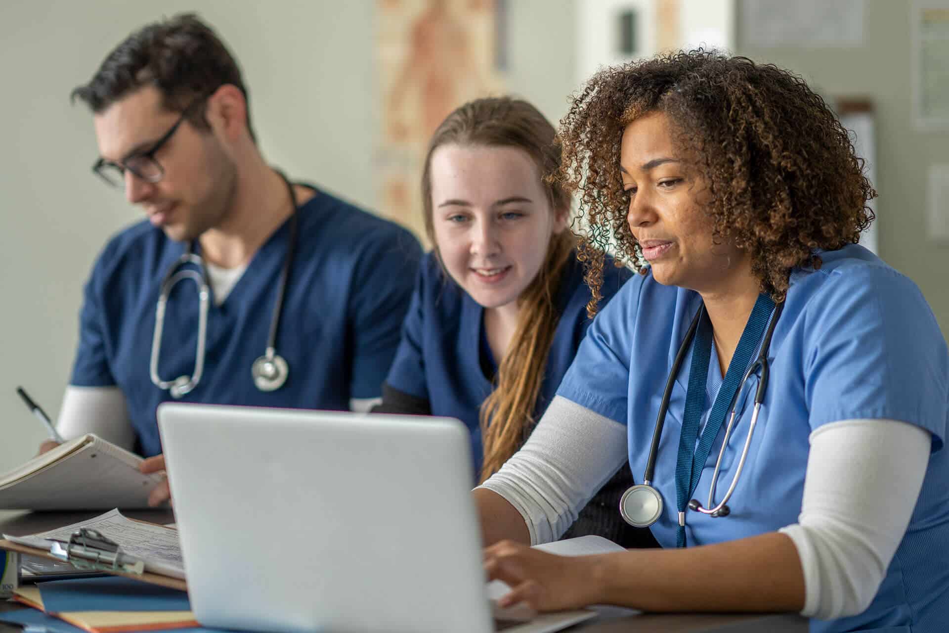Three medical professionals in scrubs are working together at a desk; two are looking at a laptop while one writes in a notebook.