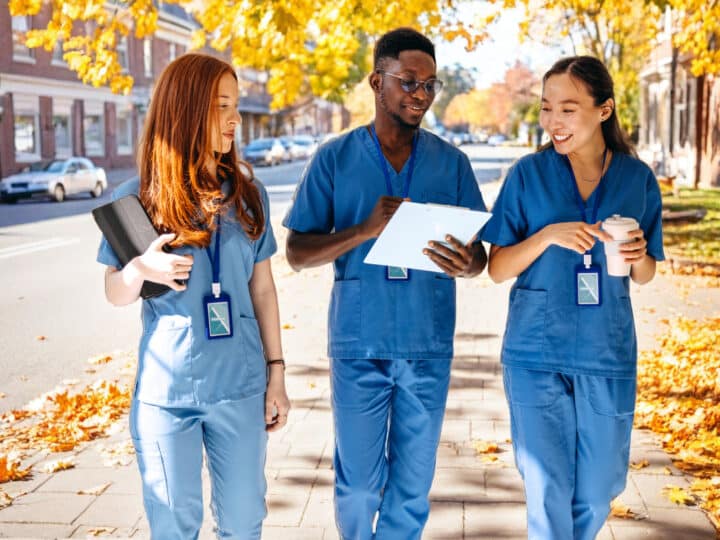 Three medical students in blue scrubs walk outside on a tree-lined street with autumn leaves, showing their potential as they smile and converse while holding tablets and notebooks.