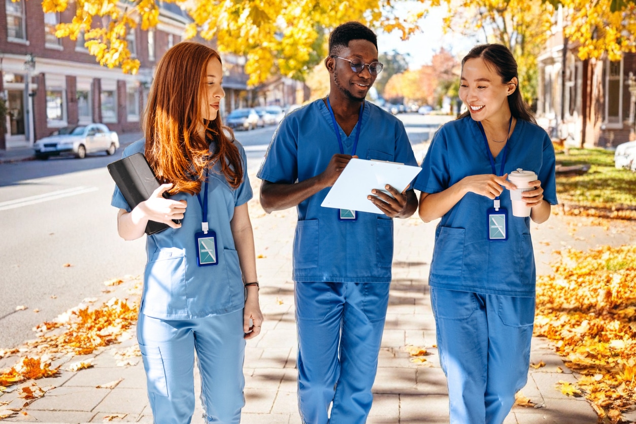 Three medical students in blue scrubs walk outside on a tree-lined street with autumn leaves, showing their potential as they smile and converse while holding tablets and notebooks.