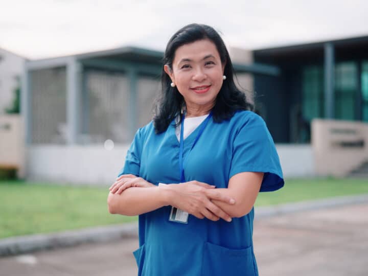 A woman wearing blue medical scrubs stands outside a building with her arms crossed, smiling at the camera, radiating confidence and potential.