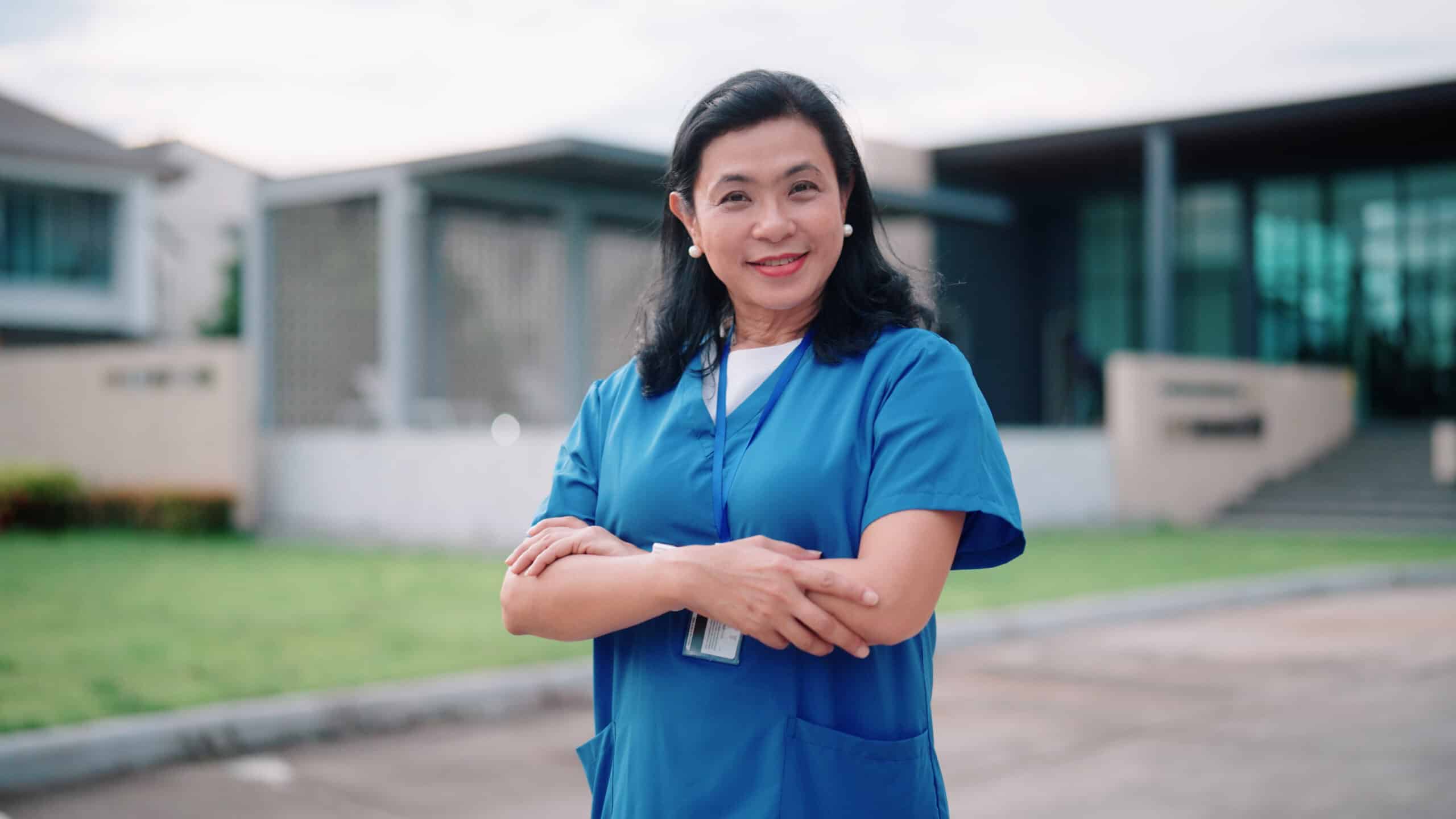 A woman wearing blue medical scrubs stands outside a building with her arms crossed, smiling at the camera, radiating confidence and potential.