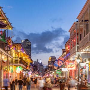 Crowds walk along a brightly lit city street lined with colorful buildings, neon signs, flags, and ads for exclusive travel nurse jobs at dusk.