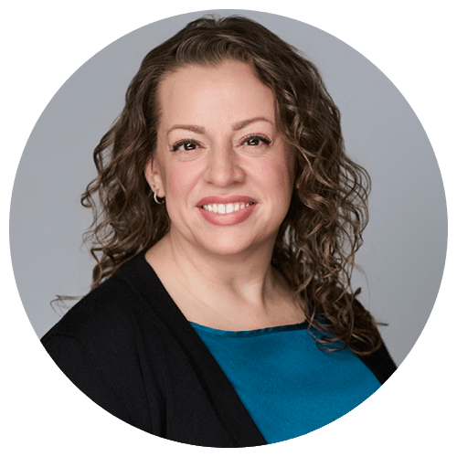A woman with wavy brown hair, wearing a teal top and black cardigan, smiles at the camera against a plain gray background.