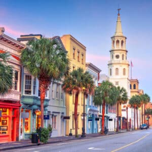 Colorful historic buildings and palm trees line a street with a white church steeple, inviting exclusive travel nurse jobs under a vibrant blue and pink sky.