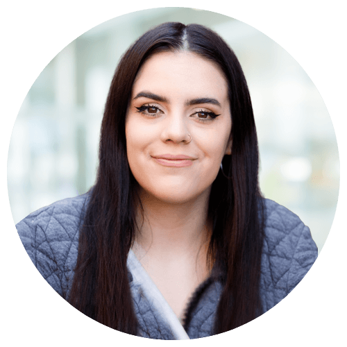 A woman with long dark hair and a grey quilted jacket is facing the camera, smiling slightly, against a blurred light background.