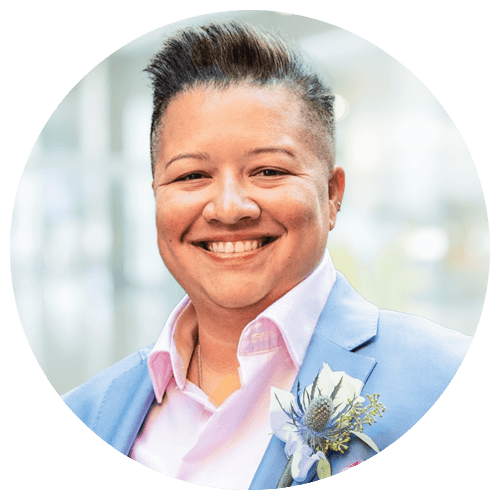 Person wearing a light blue suit jacket, pink shirt, and boutonnière, smiling at the camera with a blurred background.