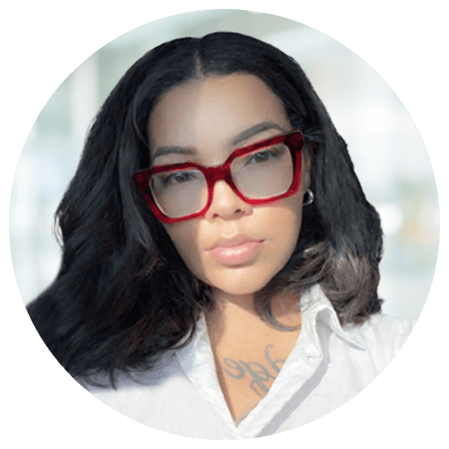 Woman with long dark hair and red glasses, wearing a white shirt, looking at the camera. Sunlight casts a shadow across the left side of her face.