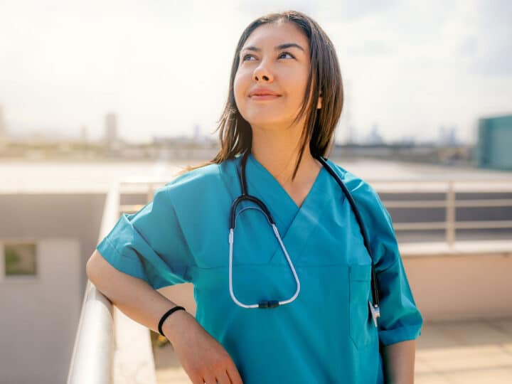 A healthcare worker in blue scrubs with a stethoscope around her neck stands outside on a rooftop, smiling—ready for new spring travel nursing assignments.