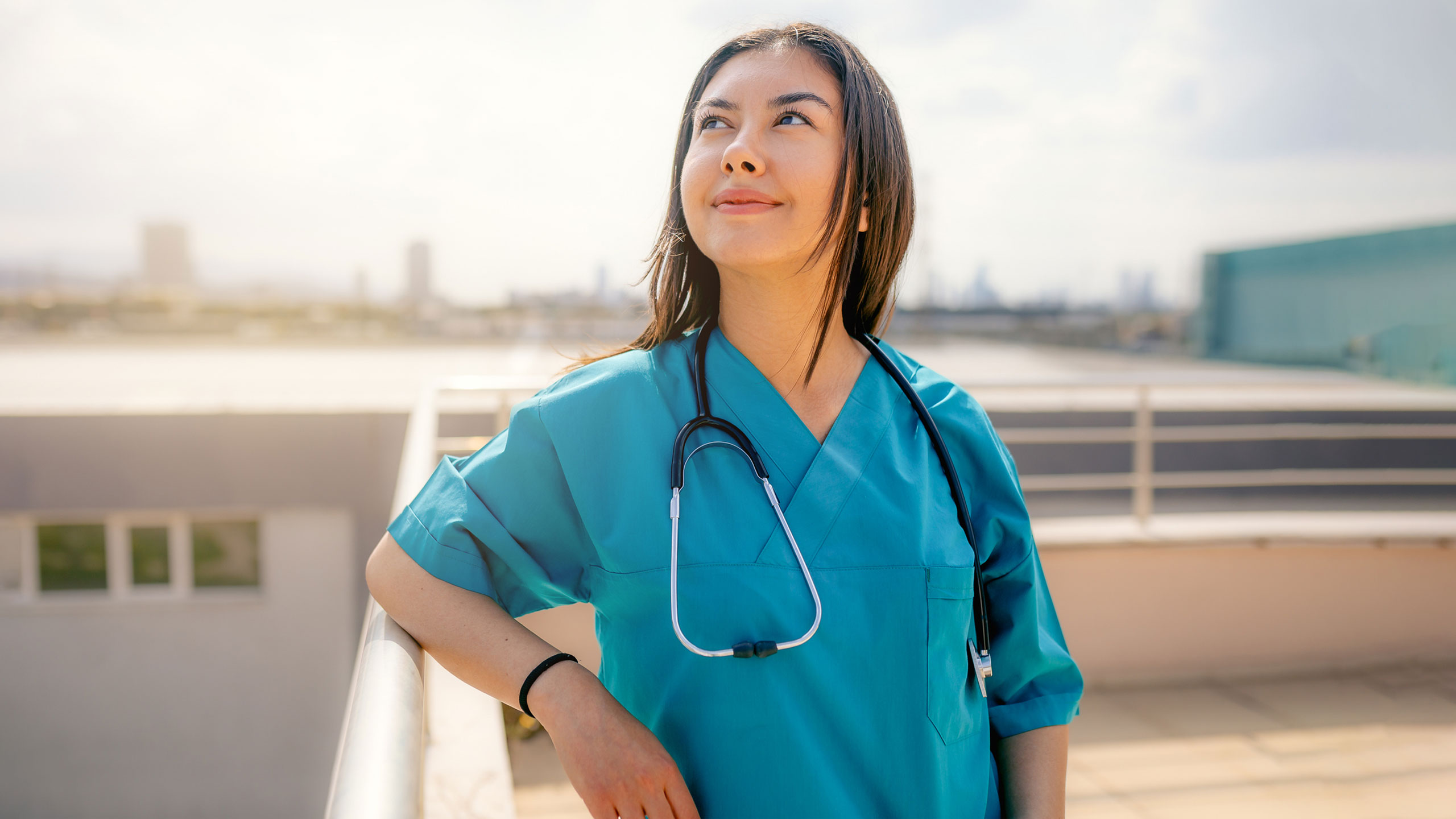 A healthcare worker in blue scrubs with a stethoscope around her neck stands outside on a rooftop, smiling—ready for new spring travel nursing assignments.