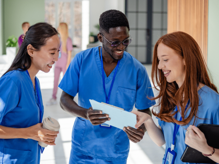 Three healthcare professionals in blue scrubs stand together in a hallway, smiling and discussing spring travel nursing assignments on a clipboard.