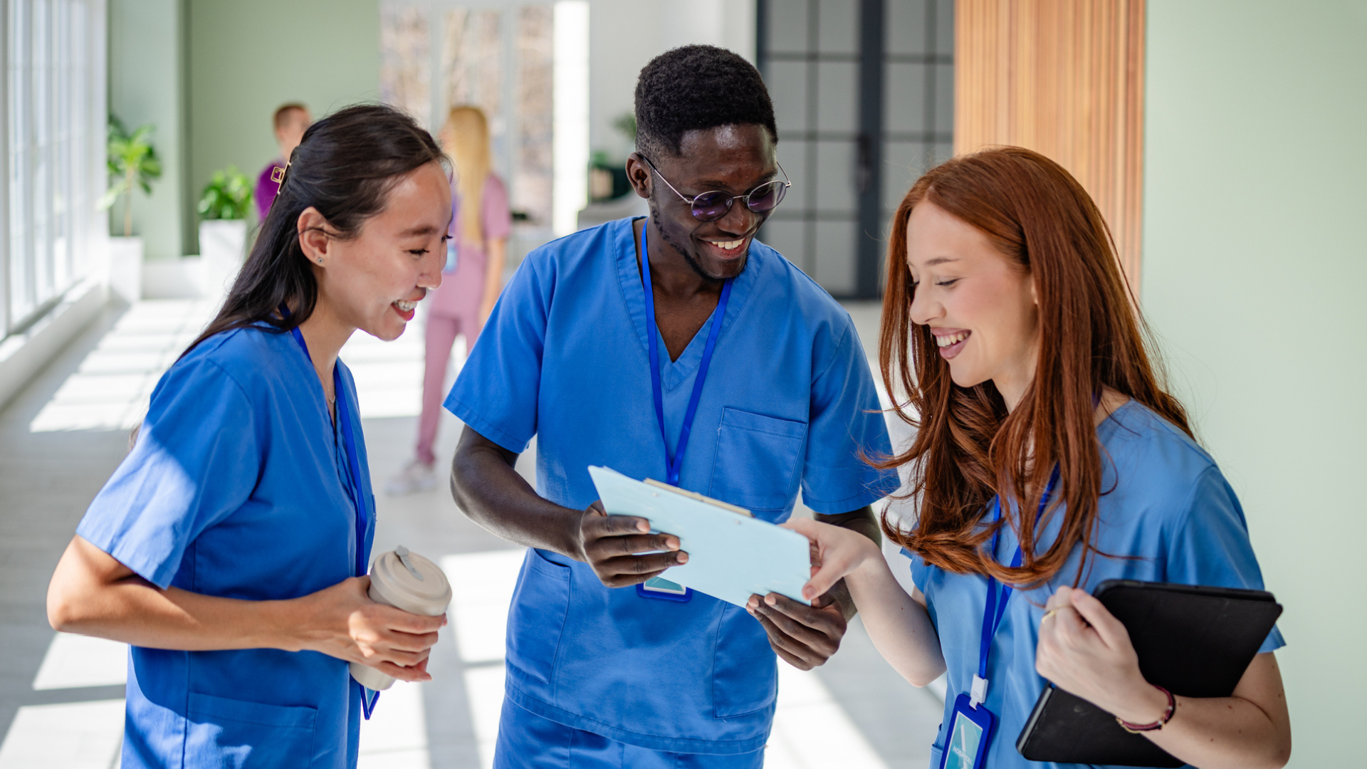 Three healthcare professionals in blue scrubs stand together in a hallway, smiling and discussing spring travel nursing assignments on a clipboard.
