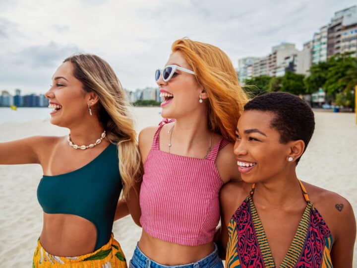 Three women stand close together, smiling and looking to the left on a sandy beach, chatting about nursing freebies & discount offers as buildings and trees line the background.