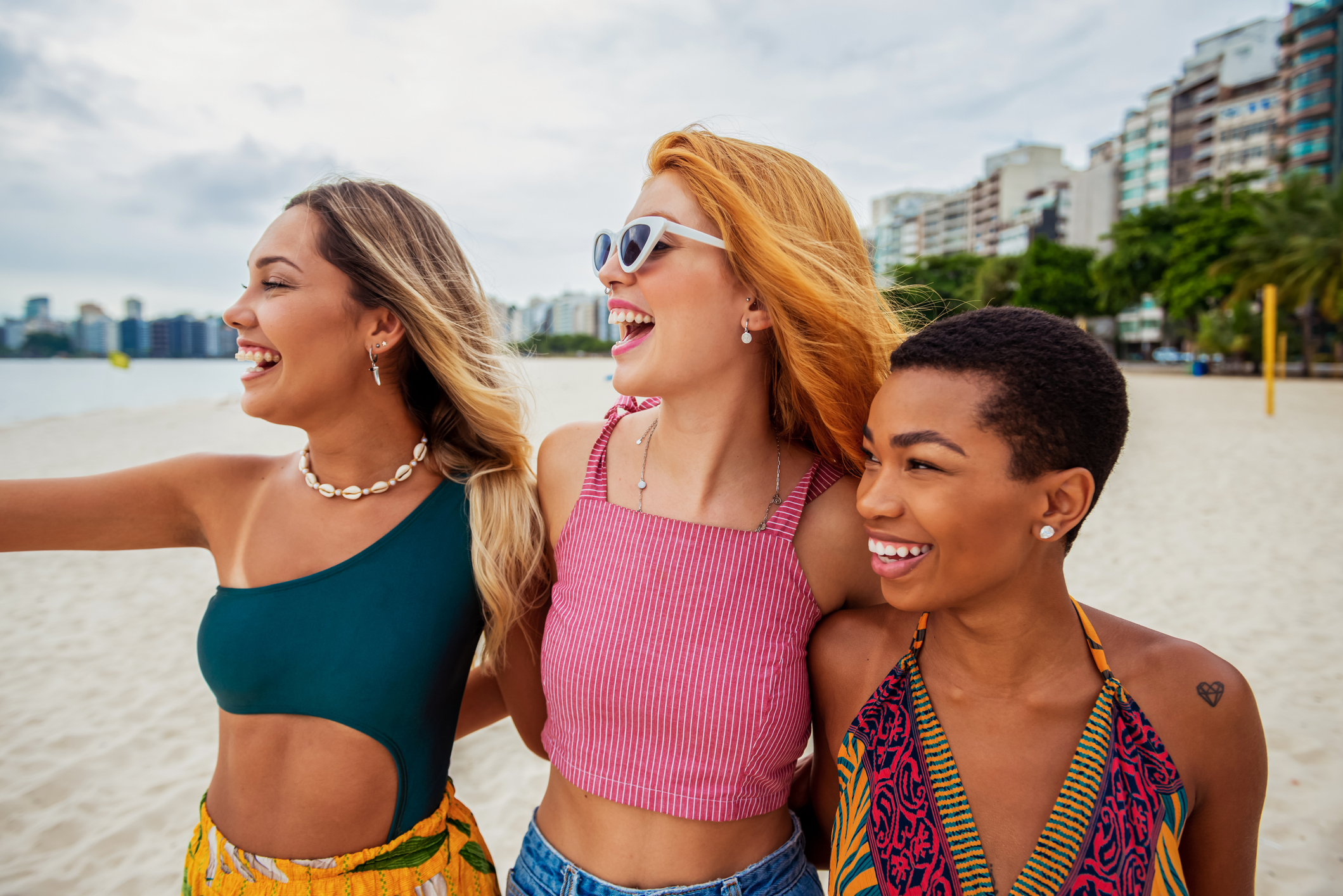 Three women stand close together, smiling and looking to the left on a sandy beach, chatting about nursing freebies & discount offers as buildings and trees line the background.