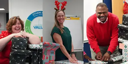 Three coworkers smile while participating in a holiday gift-wrapping activity at the office.