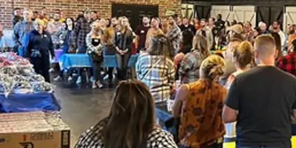 Crowd gathered around tables displaying items during an indoor community event.