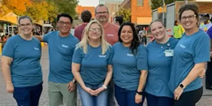 Group of seven people in matching blue shirts posing together at an outdoor community event.