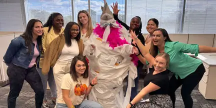 Group of eight people posing around a person wrapped in white tissue paper and wearing a bright pink scarf and pointed hat in an office.
