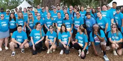 Large group of people wearing matching bright blue shirts gathered outdoors among trees and event tents.