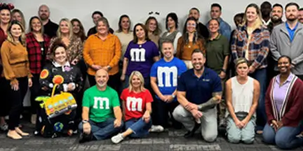 Large group posing indoors, with several people wearing colorful M&M-themed shirts and one person in a candy-themed costume.